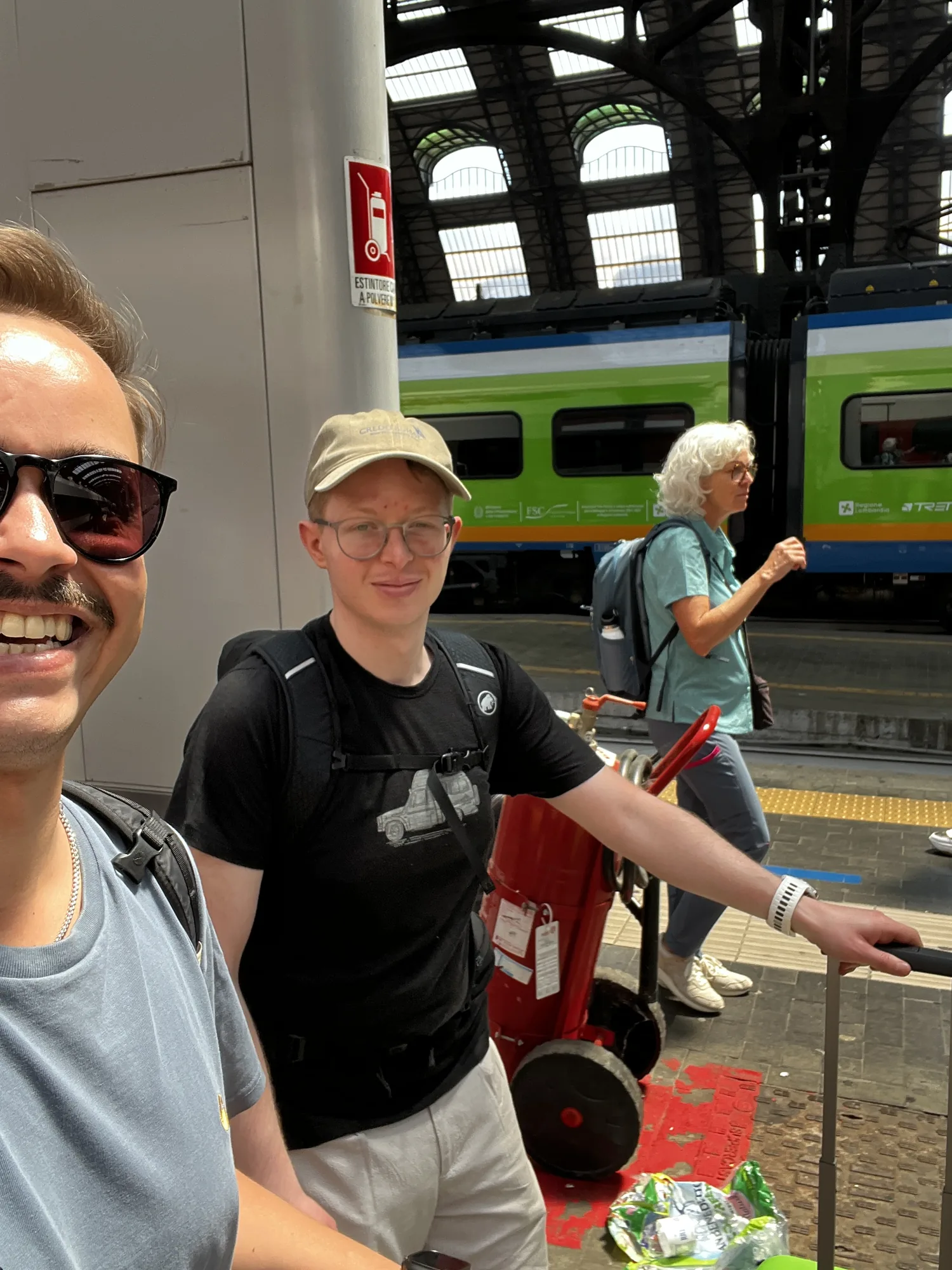Zwei Freunde am Bahnhof Milano Centrale vor einem grünen Trenitalia-Zug